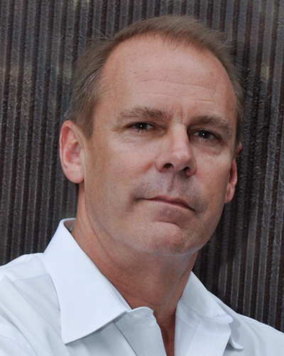 Charles Knight in an open white shirt, against a ribbed metal wall.