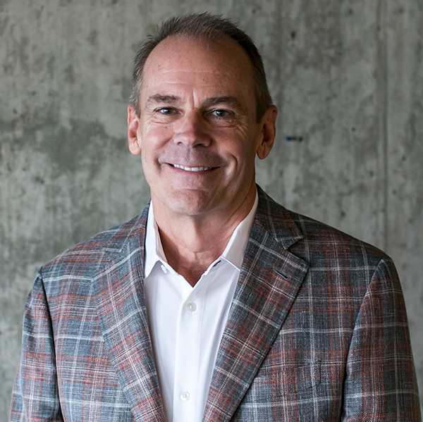 Charles Knight in a grey and rust plaid jacket over an open white collar, against a concrete wall.