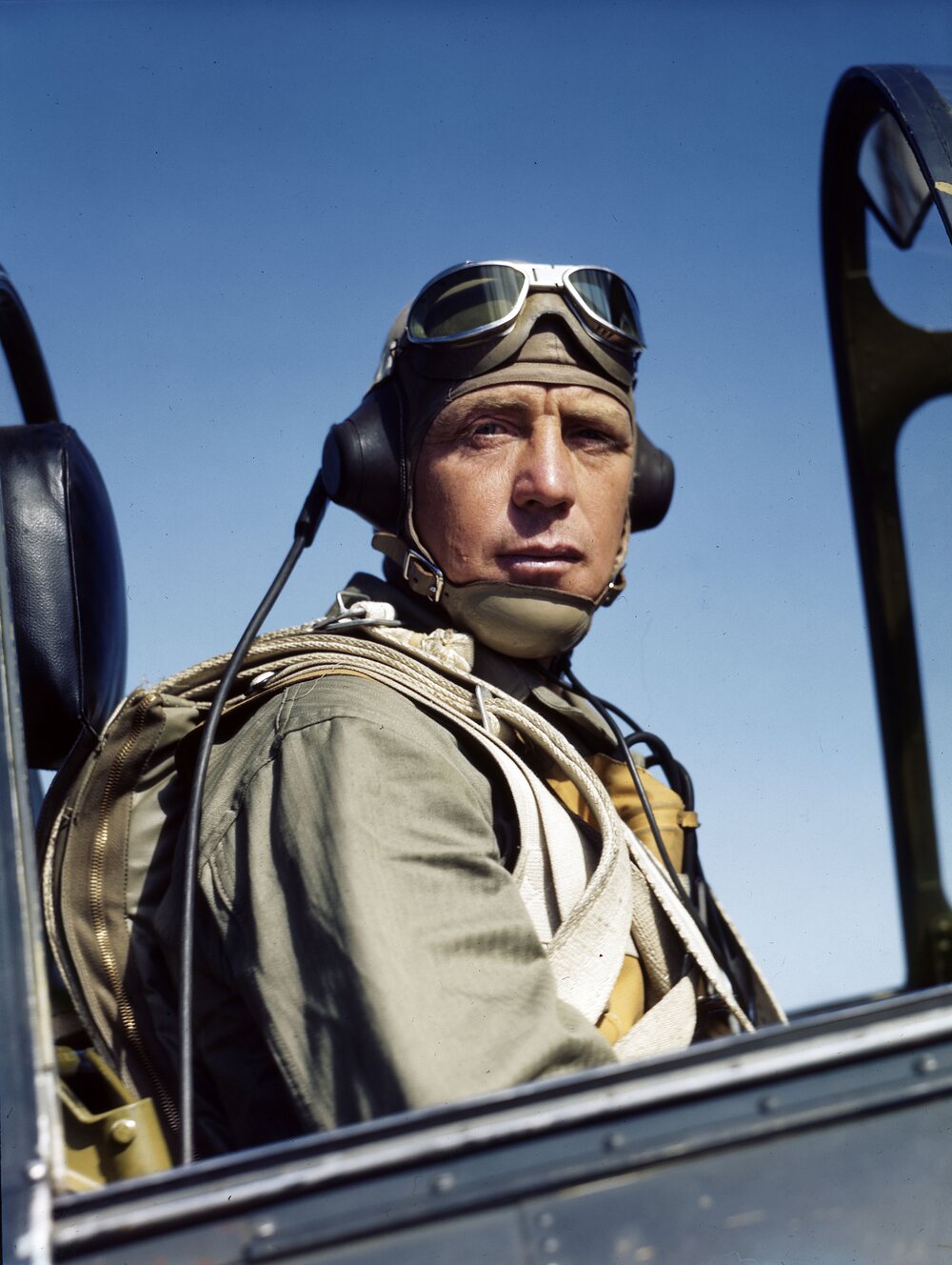 Colour photograph of a pilot in the open cockpit of a fighter, leather helmet and goggles pushed up, parachute harness over his shoulders, looking directly at the camera against a deep blue sky.