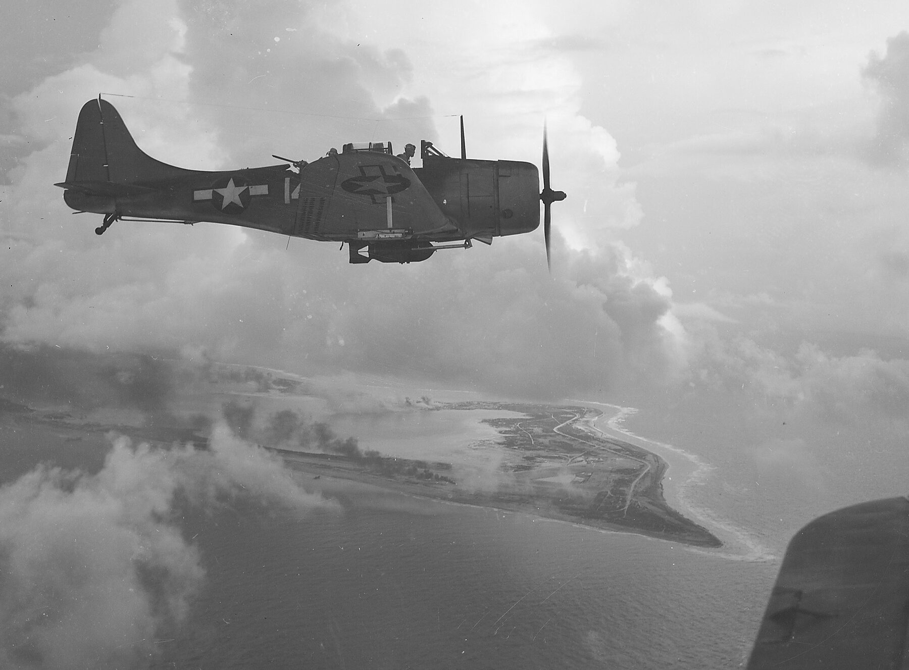 A Douglas Dauntless dive bomber banking through broken cloud, high above a low sandy island ringed by surf and streaked with smoke.