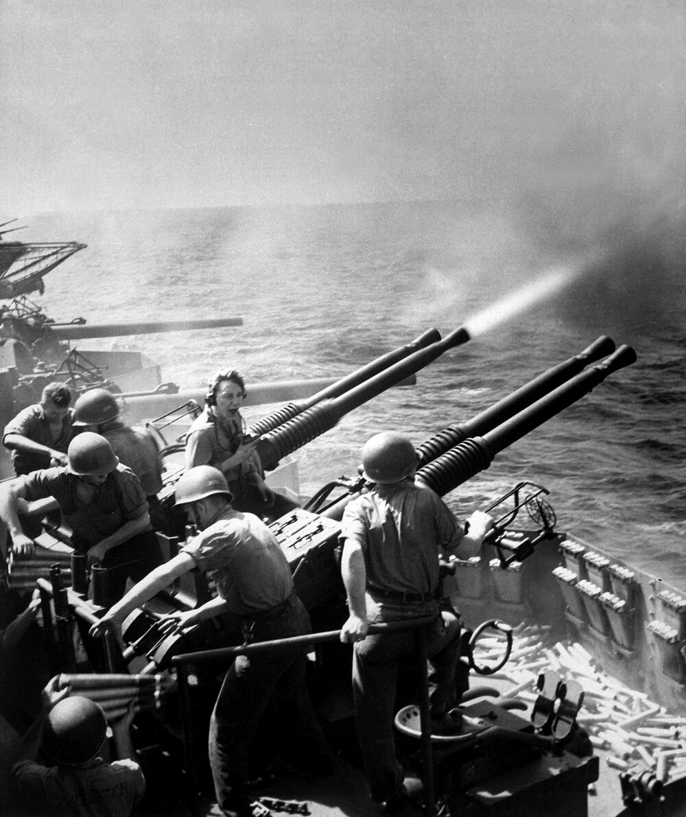 A twin 40mm anti-aircraft mount firing from a carrier's gun gallery, tracer smoke streaming from the muzzles, gun crew in helmets working the mount, spent shells across the deck.