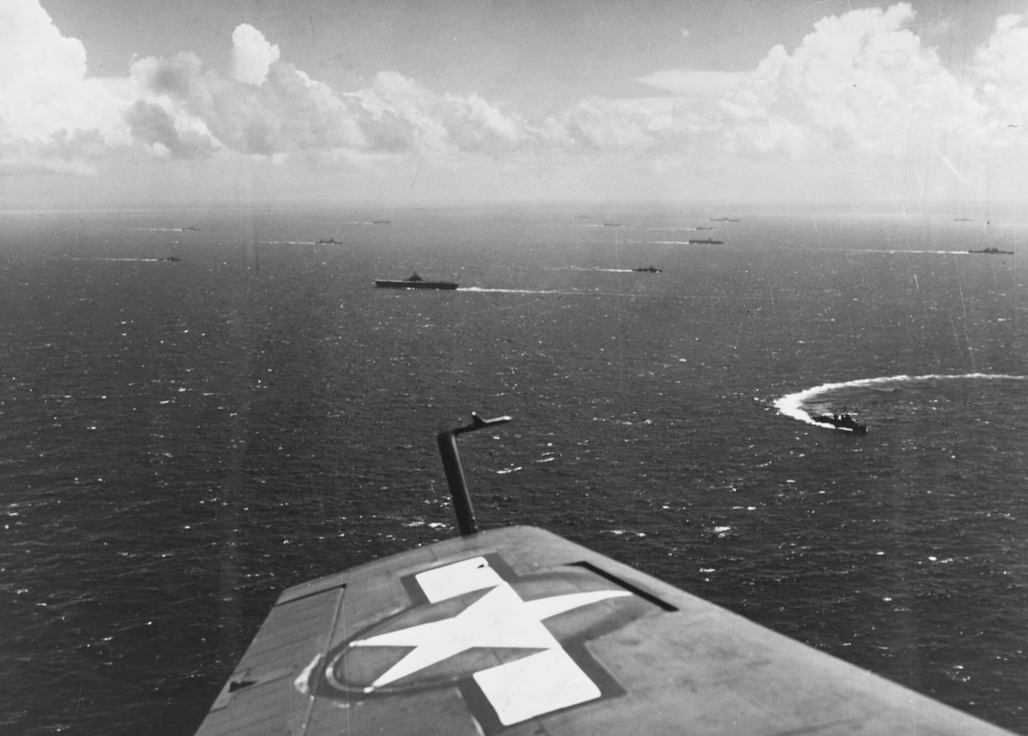 An aerial view over an aircraft wing marked with a white star: a carrier task force spread across open ocean below, ships trailing wakes toward the horizon.