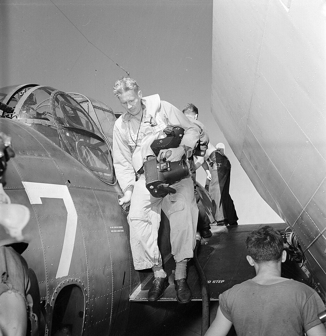 A fair-haired man in a flight suit climbs down from the wing of a Navy aircraft onto the deck, two boxy cameras slung across his chest, sailors around him.