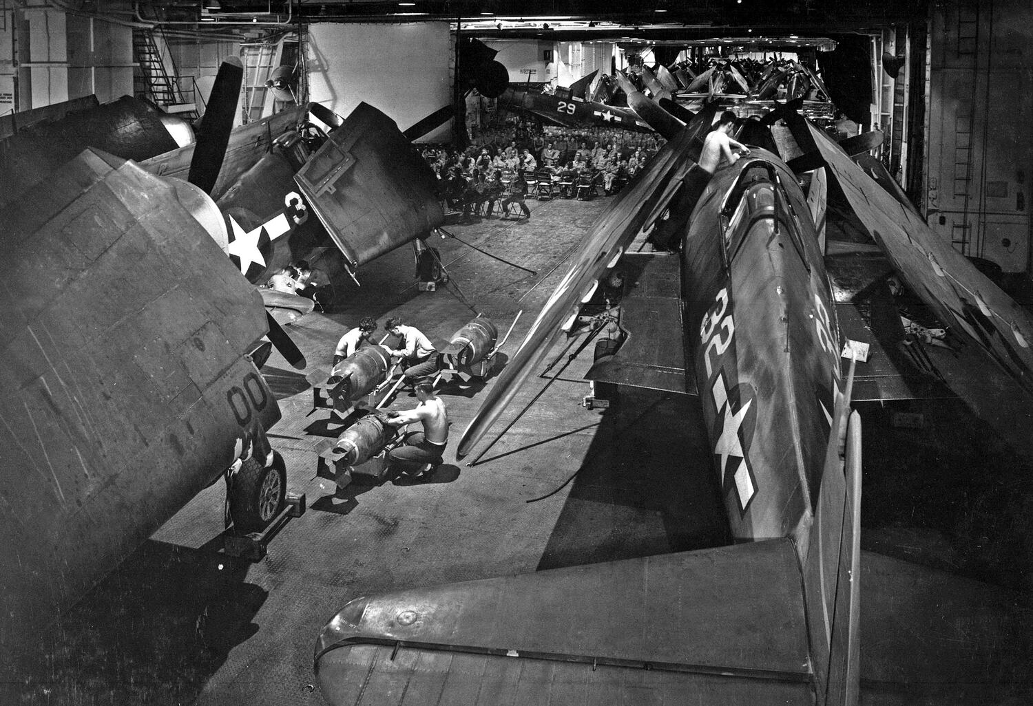 The hangar deck of an aircraft carrier, crowded with folded-wing fighters. In the foreground three men crouch over bombs on trolleys; in the middle distance, rows of seated sailors watch a film.