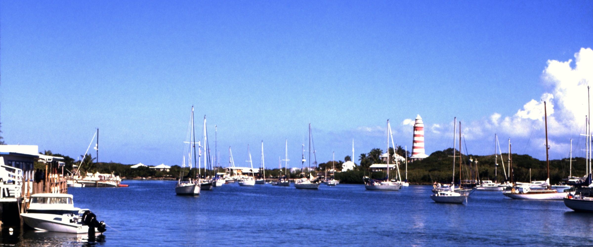 Hope Town harbour on Elbow Cay — sailing boats at anchor in flat blue water, a low green shoreline behind them, and the red-and-white striped Elbow Reef lighthouse standing on the point.